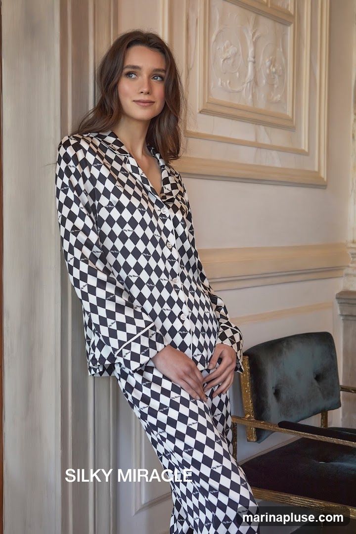 A woman with long brown hair stands leaning against a wall, wearing a black and white diamond pattern pajama set. The pajamas have a white piped trim and a subtle repeating logo pattern. She is looking off to the side with a gentle smile. In the background, a plush green velvet chair with ornate gold trim is visible. The wall behind her features decorative molding. The text "SILKY MIRACLE" is overlaid on the lower left of the image.