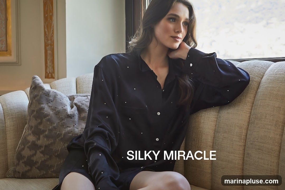 A woman with long dark hair is sitting on a couch, looking out a window. She is wearing a black silk pajama set adorned with small, scattered rhinestones. The couch is upholstered in a textured beige fabric, and a decorative gray pillow rests beside her. The background features a wall with ornate molding and a framed artwork. The text "SILKY MIRACLE" is overlaid on the image.