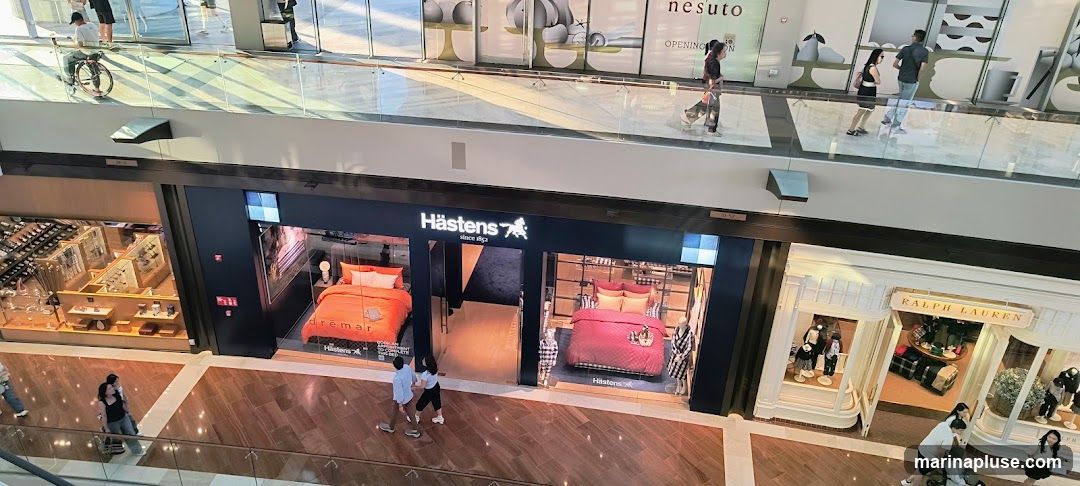 An overhead view of a multi-level department store interior. On the lower level, a Hästens store displays beds with vibrant bedding. To the right, a Ralph Lauren store showcases clothing and accessories. Other shops with display cases are visible on the left and in the background. People are seen browsing and walking through the mall.