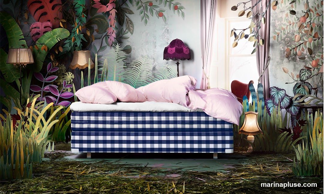 A bed with pink bedding is set against a backdrop of lush, stylized foliage and a whimsical mural. The bed frame is covered in a blue and white gingham fabric. Several lamps are placed around the scene, some with ornate shades. The floor is covered in a layer of grass and twigs, creating an enchanted forest atmosphere.