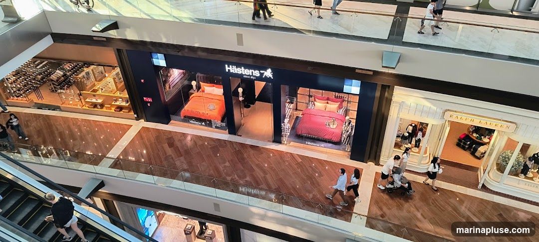 An overhead view of a brightly lit, multi-level department store interior. In the foreground, people are seen on an escalator. Further back, several shopfronts are visible, including Hästens, displaying beds, and Ralph Lauren, showcasing clothing. Other shops with various merchandise are also present.