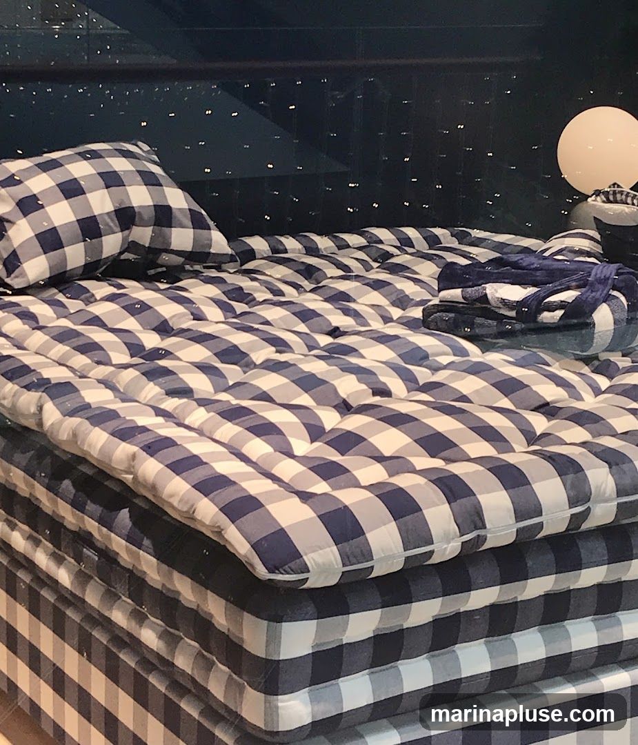 A display of stacked mattresses and bedding with a blue and white gingham pattern. A pillow rests on the top mattress, and folded blankets are placed at the foot of the bed. The background is dark with small, bright lights, and a round lamp is visible to the right.