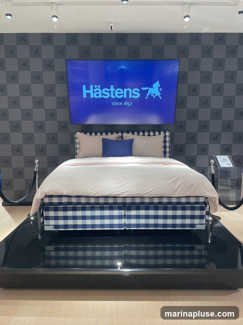 A Hästens bed is displayed in a showroom. The bed has a blue and white checkered base and is covered with light pink bedding and two pillows. Behind the bed, a large screen displays the Hästens logo and the text "since 1852". The wall behind the screen has a repeating pattern of horses. The display is roped off with stanchions.
