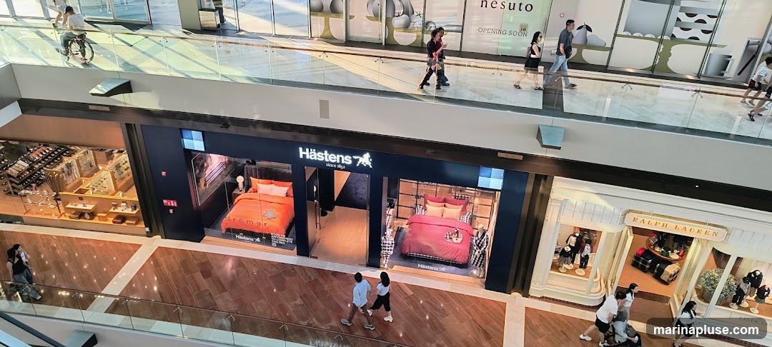 An overhead view of a shopping mall interior, showcasing the storefronts of Hästens and Ralph Lauren. The Hästens store features displays of beds with vibrant bedding. Adjacent to it, the Ralph Lauren store displays clothing on mannequins and shelves. People are seen walking on the mall concourse.