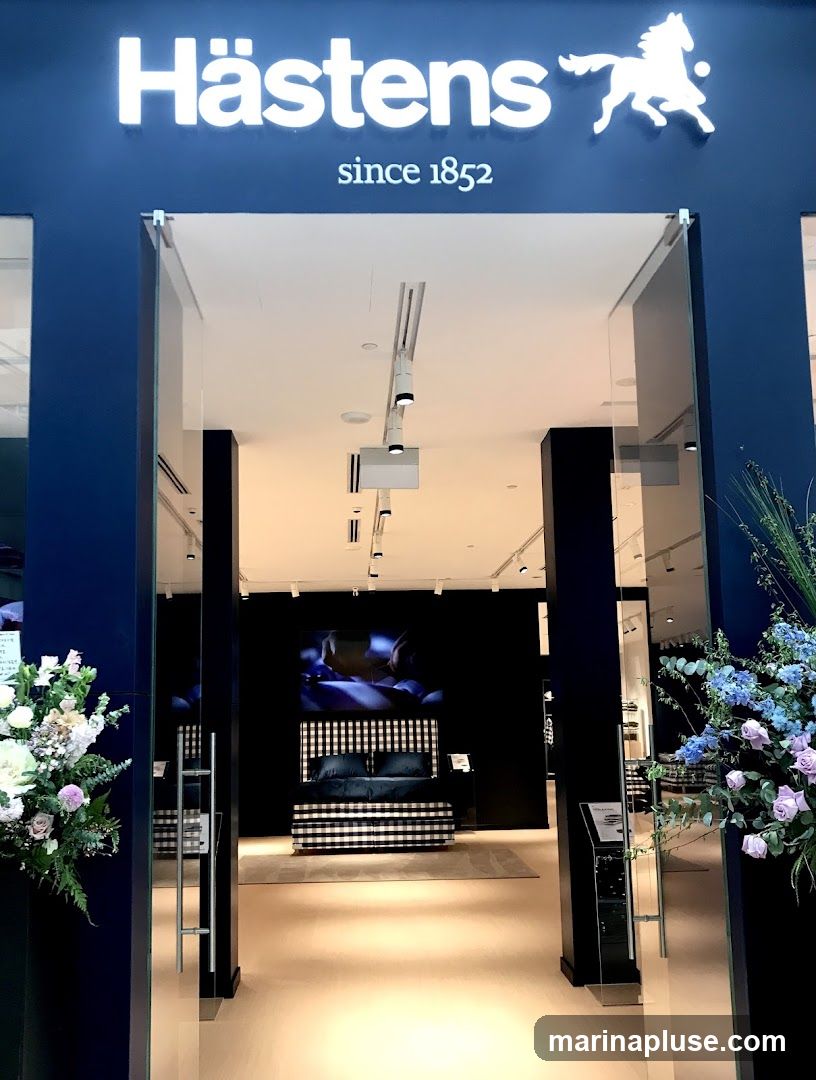 The entrance to a Hästens store, featuring the brand's name and logo above the doorway. The store interior is visible, showcasing a bed with a black and white checkered headboard and bedding. Floral arrangements are placed on either side of the entrance.