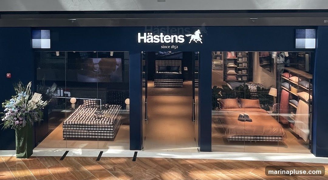 The exterior of a Hästens store, featuring large glass windows displaying beds and bedding. The store's name and logo are visible above the entrance. Inside, beds are set up with various bedding styles, and shelves display pillows and other linens. A floral arrangement is placed to the left of the entrance.