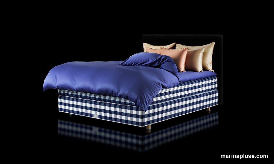 A bed with a black headboard is dressed in a deep blue duvet and several decorative pillows in shades of peach and gold. The base of the bed is covered in a navy and white gingham fabric. The bed is presented against a black background with a reflective surface below.