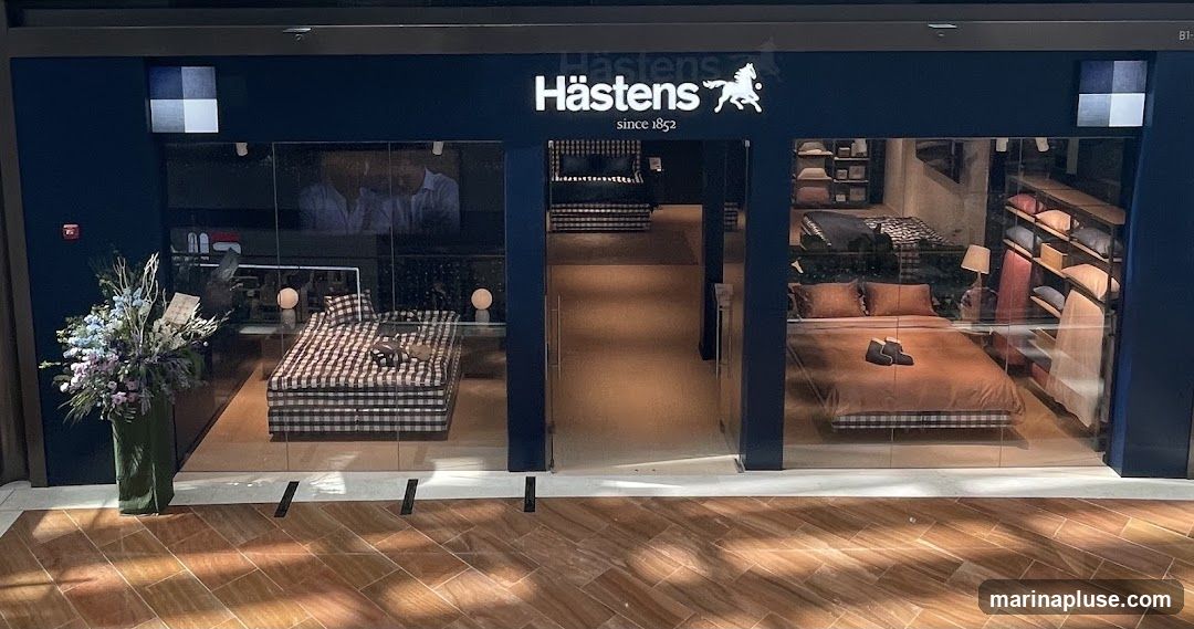 The storefront of a Hästens store, featuring large glass windows displaying beds and bedding. The store's name and logo are visible above the entrance. Inside, beds are set up with various bedding options, and shelves display pillows and other accessories. A floral arrangement is placed to the left of the entrance.