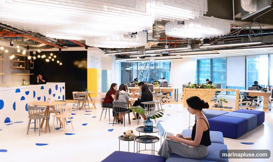 An interior view of a bright and modern co-working space with various seating and work areas. In the foreground, a woman is working on a laptop while seated on a blue modular sofa. In the mid-ground, several people are seated at tables, some in discussion, others working. The space features a counter area with a person behind it, and a yellow and blue locker area. The ceiling has exposed pipes and a decorative lighting installation. Large windows offer a view of a city skyline.
