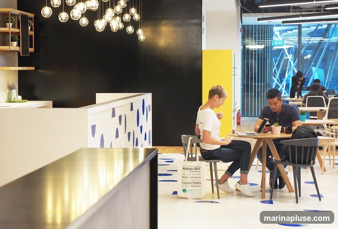 A bright and modern co-working space features a reception desk with decorative blue patterns. A woman in a white t-shirt and jeans sits at a round table, looking at her laptop. A man in a dark polo shirt sits opposite her, looking at his phone. In the background, other individuals are working at tables, and large windows offer a view of the city. A yellow partition adds a pop of color to the space. A tote bag with text listing various locations is placed on the floor near the woman.
