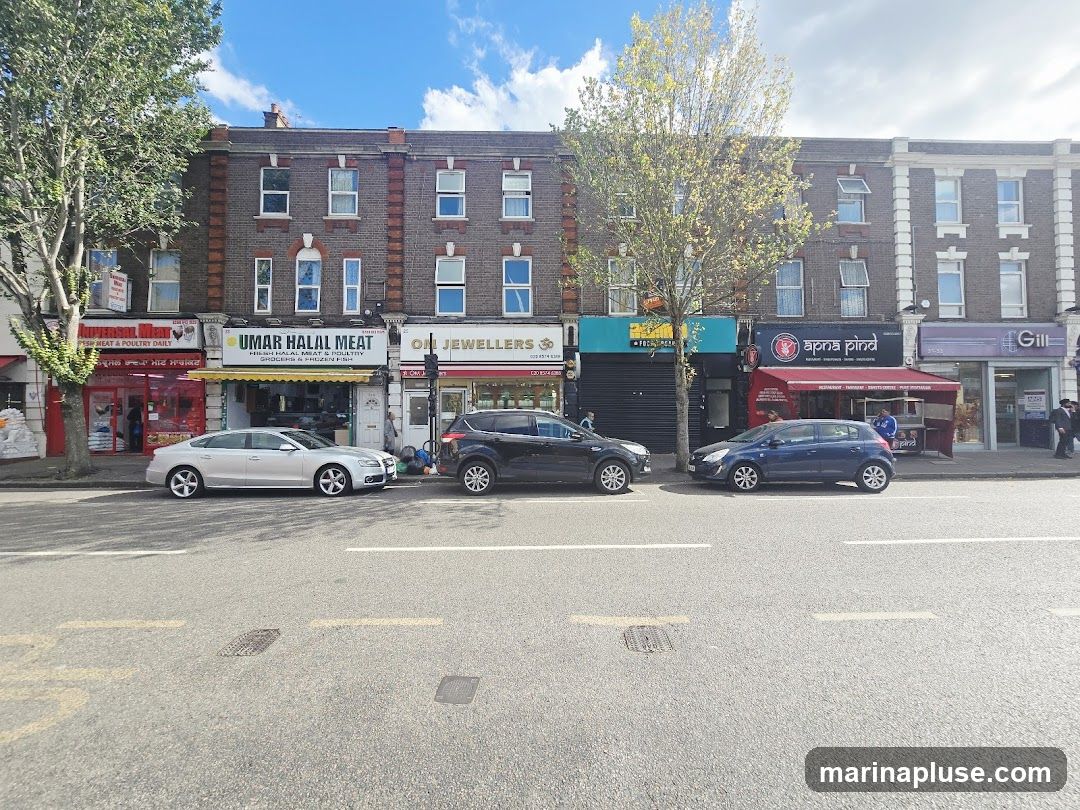 A row of shops with various businesses, including a halal meat shop, a jeweler, and a restaurant. Cars are parked along the street in front of the shops.
