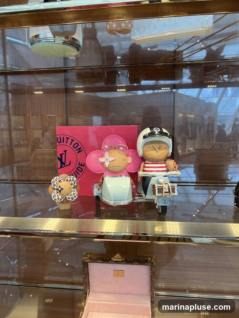 A display case with Louis Vuitton merchandise. In the center, there are two decorative figures, one resembling a flower with sunglasses and the other a person on a scooter wearing a helmet. To the left is a pink box with the Louis Vuitton logo. Below, an open decorative box with a pink interior is displayed.