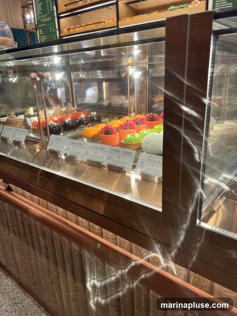 A display case filled with various colorful pastries and cakes. Small white cards with names and prices are placed in front of each item. The background shows shelves with baked goods.