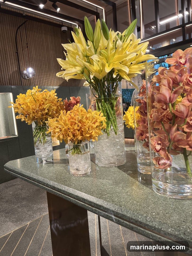 A close-up shot of a display of flowers in glass vases on a speckled countertop. The flowers include yellow lilies, yellow orchids, and peach-colored orchids. The background shows a wooden paneled wall with lighting fixtures and a security camera.