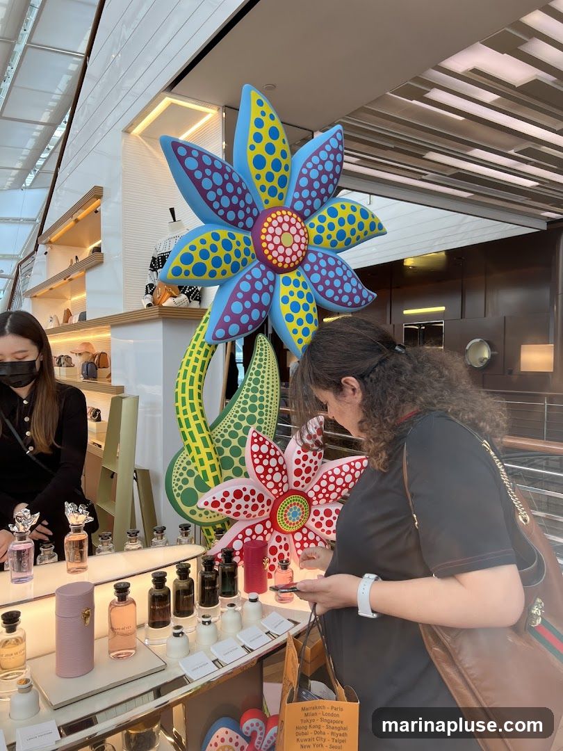 A display of perfume bottles on shelves in a store, with a large, colorful flower art installation in the foreground. Two women are looking at the perfumes. A sign with city names is visible at the bottom right.