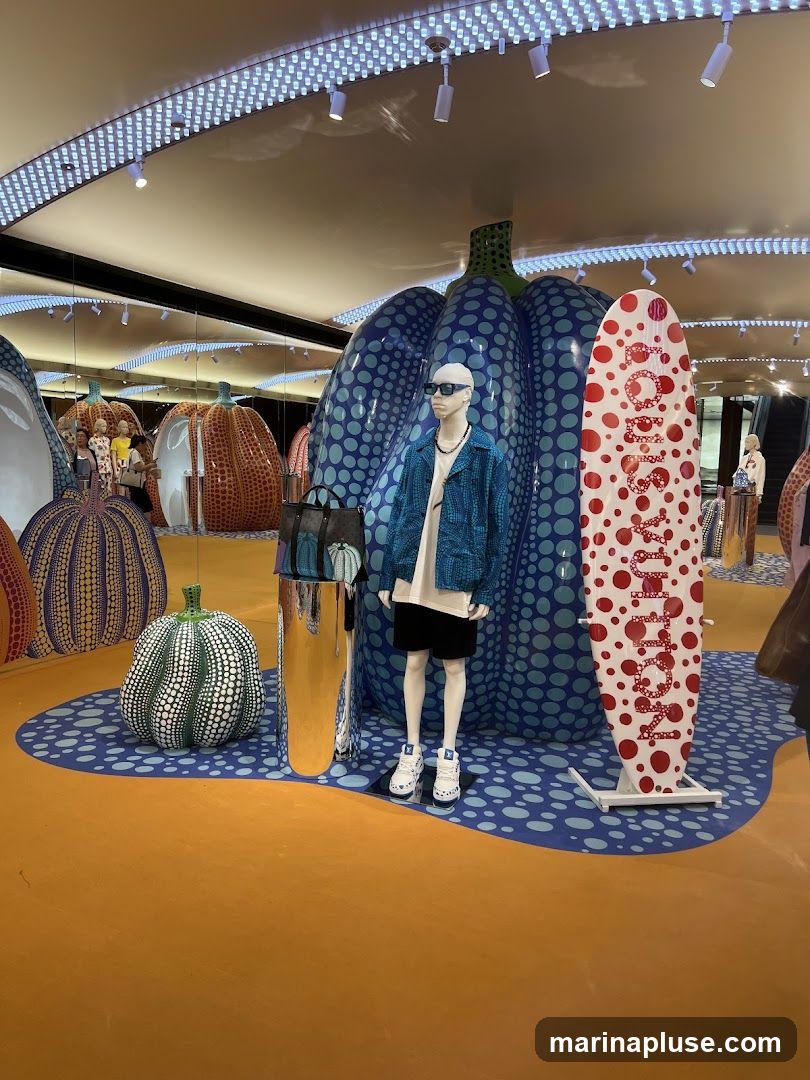 A store interior features a display with large, polka-dotted pumpkin sculptures in various colors, including blue, orange, and green. A mannequin dressed in a blue patterned jacket, white t-shirt, and black shorts stands in front of a large blue pumpkin. Next to the mannequin is a white surfboard decorated with red polka dots and the word "LOUIS VUITTON" in red lettering. The floor is orange with a blue and white polka dot pattern. Mirrors on the back wall reflect the display and other mannequins. The ceiling has a row of bright white lights.