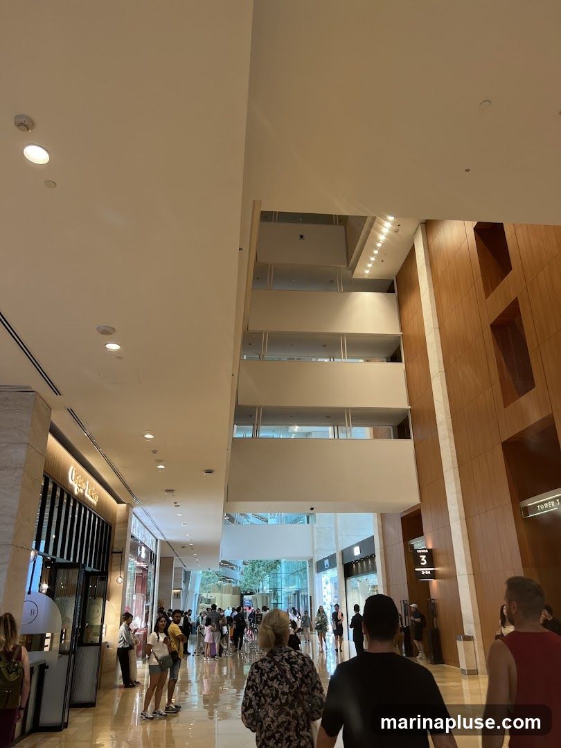 A wide shot of the interior of a modern shopping mall with a high ceiling and multiple levels. People are walking along a polished floor. On the left, there are several shops with illuminated signs. One sign reads 'Grand Palais'. On the right, a wooden-paneled wall features signage for 'Tower 3'.