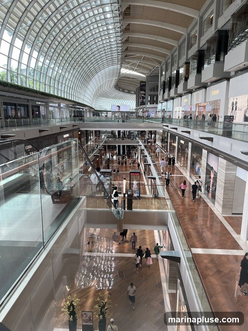 A high-angle view of the interior of a large, multi-level shopping mall. The architecture features a distinctive curved glass roof and multiple levels of shops and walkways. People are seen browsing and walking on the different floors. The mall has a clean, contemporary design with polished floors and well-lit storefronts.