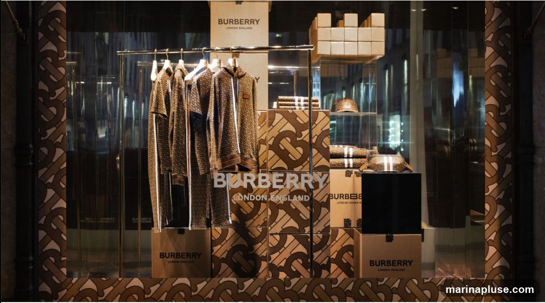 A store window display featuring Burberry clothing and accessories. The display includes several garments on hangers, such as track jackets and t-shirts, all with the Burberry logo pattern. There are also folded items, hats, and boxes with the Burberry branding. The background is dark and reflective, showing the interior of the store.
