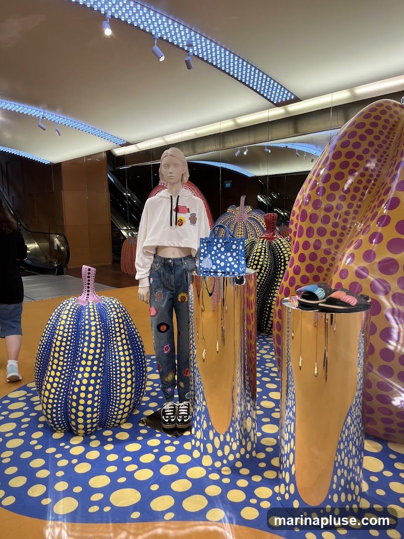 A retail display features a mannequin dressed in a white cropped hoodie and jeans adorned with colorful patches. The mannequin stands amidst large, polka-dotted sculptures resembling pumpkins and other organic shapes in vibrant yellow, purple, and blue. Two reflective cylindrical pedestals display a blue handbag and a pair of sandals. The floor is covered in a blue and yellow polka-dot pattern, and an escalator is visible in the background.