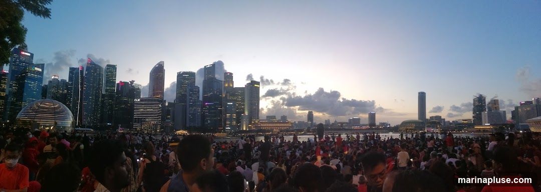 A panoramic view of a city skyline at dusk, with numerous skyscrapers illuminated. A large crowd of people are gathered in the foreground, facing the skyline. The sky is a gradient of blue and orange, with some clouds.