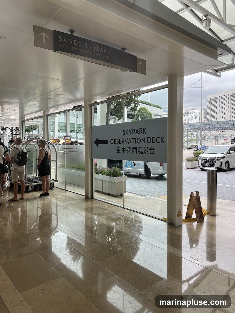 A sign indicating directions to the Sands Skypark and Skypark Observation Deck. The sign is in English and Chinese. People are visible in the background near a luggage cart. Outside, a white car and buildings can be seen.