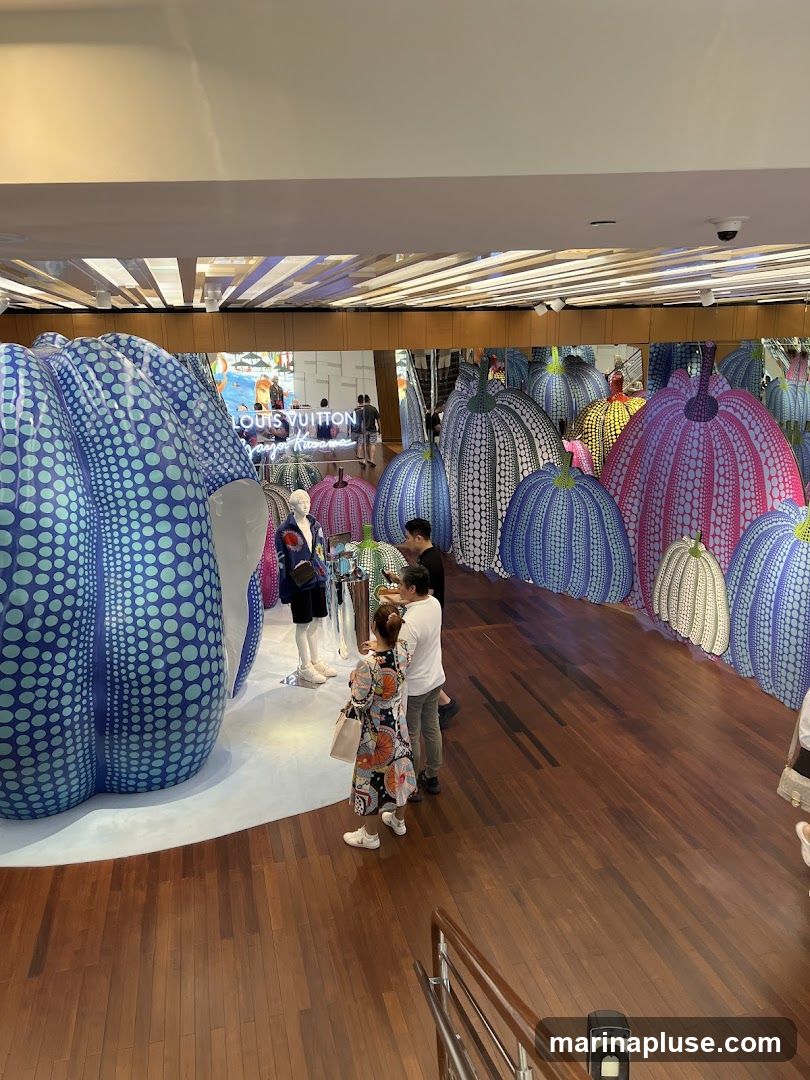 An interior view of a Louis Vuitton store featuring a large-scale art installation by Yayoi Kusama. The installation consists of numerous large, inflatable pumpkin sculptures in various colors, including blue, pink, yellow, and white, all adorned with Kusama's signature polka dots. Several mannequins are visible, dressed in what appear to be fashion items. People are observing the artwork and browsing the store. The store's interior features a polished wooden floor and a ceiling with a modern design. A neon sign that reads "LOUIS VUITTON Yayoi Kusama" is prominently displayed in the background.