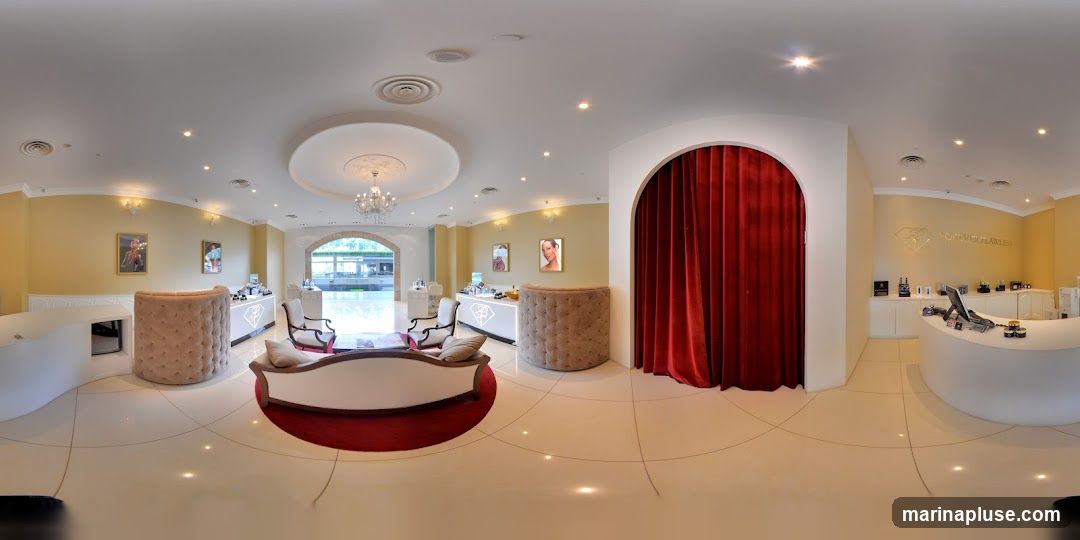 A 360-degree view of a high-end beauty store interior. The space features a reception desk with a computer, various skincare and cosmetic products displayed on shelves and counters, and elegant seating areas. The walls are painted a warm yellow, and the floor is tiled with large, light-colored squares. A large archway with red velvet curtains is visible on one side. Framed pictures of models and artwork adorn the walls. The overall ambiance is sophisticated and inviting.