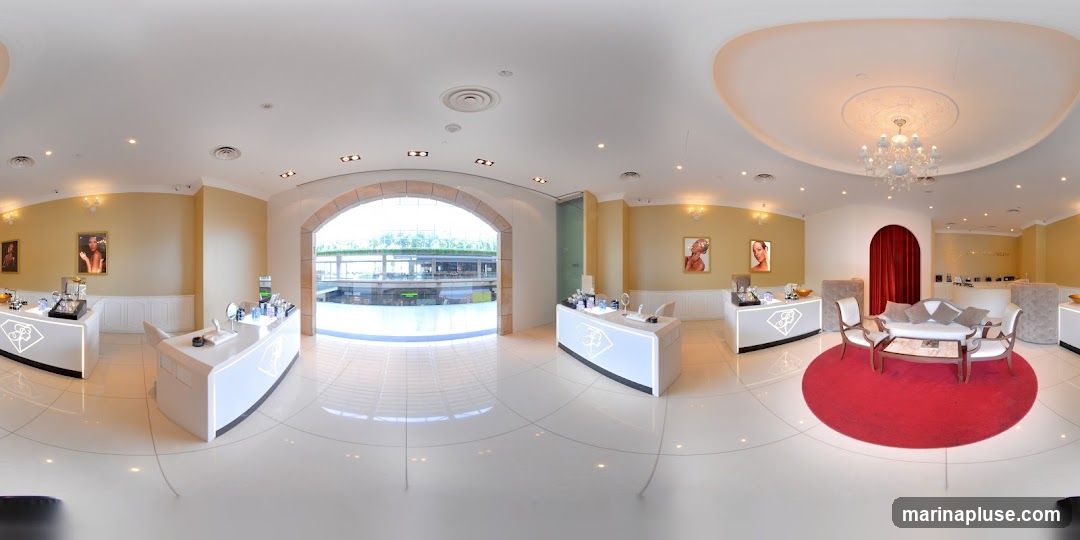 A panoramic view of a well-lit, modern jewelry store interior. The store features white display counters with illuminated logos, elegant seating areas with a red rug, and framed portraits on the walls. An arched entryway reveals a view of an outdoor shopping area.