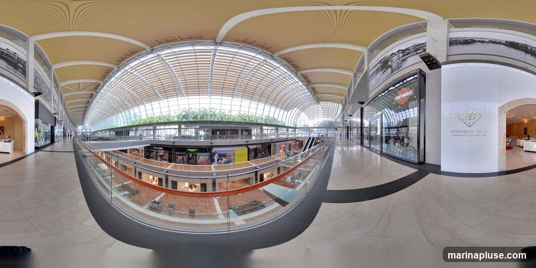 A panoramic view of the interior of a large, modern shopping mall. The architecture features a curved, glass roof that allows natural light to flood the space. Multiple levels of shops are visible, with escalators and walkways connecting them. Some shops have large display windows showcasing merchandise. One prominent storefront displays the Harley-Davidson logo. Another area features a sign for 'Forever Flawless: Diamond Infused Skin Care'. The mall appears to be clean and well-maintained, with polished floors and contemporary design elements.