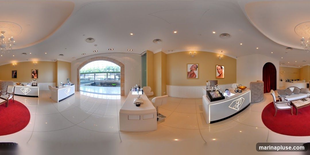 The image shows the interior of a well-lit, luxurious boutique. The walls are painted a warm yellow, and the floors are polished white tile. There are several display counters with illuminated logos, showcasing various products. Seating areas with plush sofas and chairs are arranged on red circular rugs. Artwork depicting faces adorns the walls. An arched entryway leads to an area with natural light. Chandeliers hang from the ceiling, adding to the opulent ambiance.