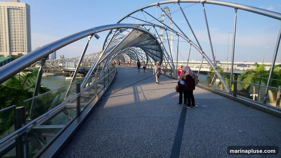 A view of the Helix Bridge in Singapore, a pedestrian bridge with a unique DNA-like structure. People are walking on the bridge under a clear blue sky. In the background, there are tall buildings and palm trees.