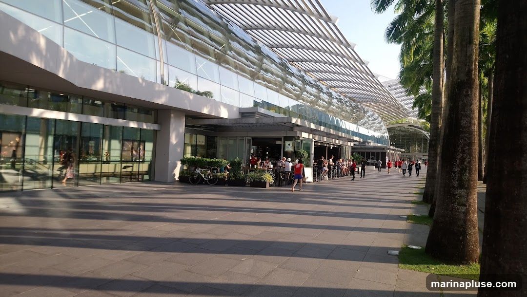 A wide paved walkway runs alongside a contemporary building with a glass facade and a distinctive slatted roof. Several palm trees line the right side of the walkway, casting long shadows. In the middle ground, an outdoor seating area for a restaurant or cafe is visible, with tables and chairs set up. People are seated at some of the tables, and others are walking along the path. Bicycles are parked near the seating area.