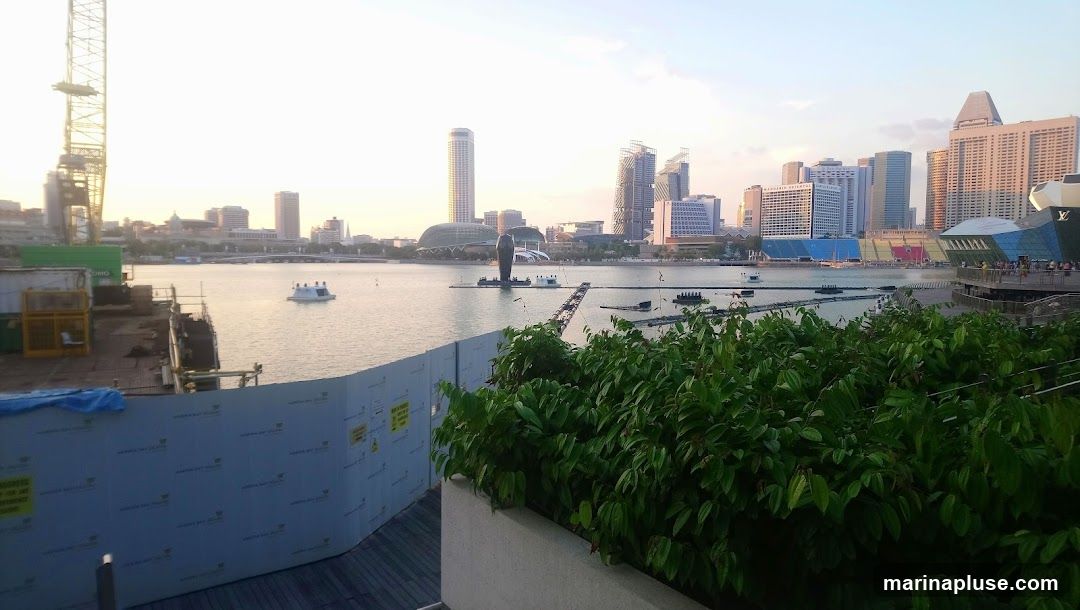A wide shot of the Marina Bay skyline at sunset. In the foreground, there is a construction site with a crane and fencing. Lush green foliage is visible in the lower right corner. The bay itself is calm, with a few boats and floating structures. The background features a dense collection of modern skyscrapers, including the Marina Bay Sands resort and the ArtScience Museum. The sky is a soft gradient of orange and yellow, indicating the end of the day.