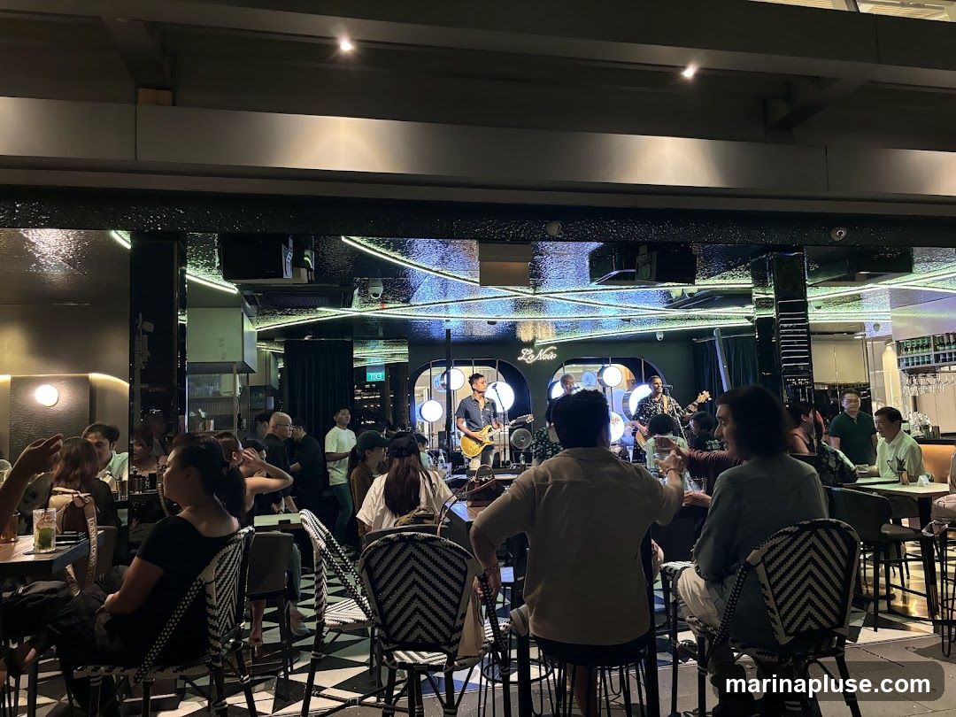A live band is performing on a stage in a dimly lit bar. Several patrons are seated at tables and chairs, facing the stage. The bar has a modern design with reflective surfaces and neon lighting.