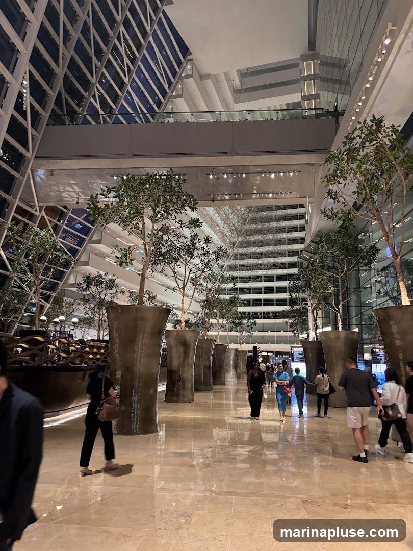 A wide shot of a modern atrium with a high, glass-paneled ceiling and multiple levels of balconies. Large, decorative planters with trees are placed throughout the polished marble floor. Several people are walking through the space.