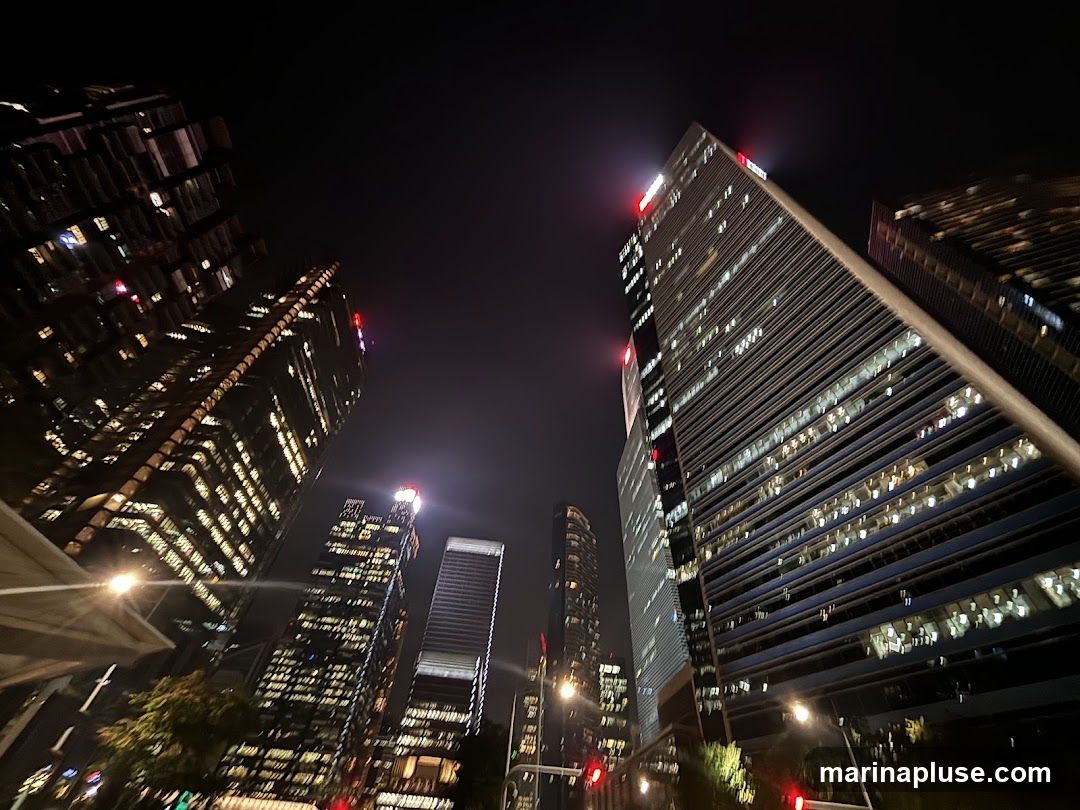 A low-angle shot of several illuminated skyscrapers against a dark night sky. The buildings have many windows, most of which are lit, suggesting occupancy. The architecture is modern, with sleek lines and reflective surfaces. A street light and traffic lights are visible in the foreground, indicating an urban environment.