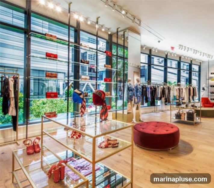A well-lit, modern retail store interior showcasing luxury fashion items. Racks of clothing, handbags on display shelves, and mannequins dressed in stylish outfits are visible. A large window offers a view of the outdoors, and a plush red ottoman sits in the center of the floor.