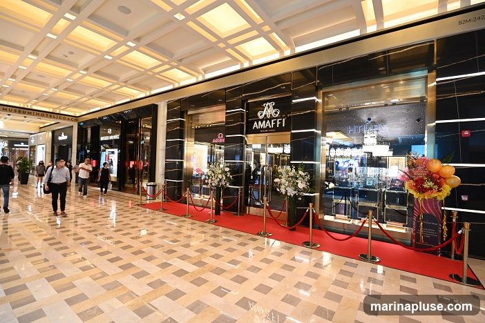 A well-lit interior of a luxury retail store with a polished marble floor and a high, coffered ceiling. The storefront features a black marble facade with gold accents. A red carpet leads to the entrance, lined with stanchions and ropes. Floral arrangements flank the entrance. The store's name, "AMaffi," is prominently displayed above the entrance. Other store names like "MUSEUM|HOTEL|COACH" and "ROLEX" are visible in the background. Several people are walking in the mall area.
