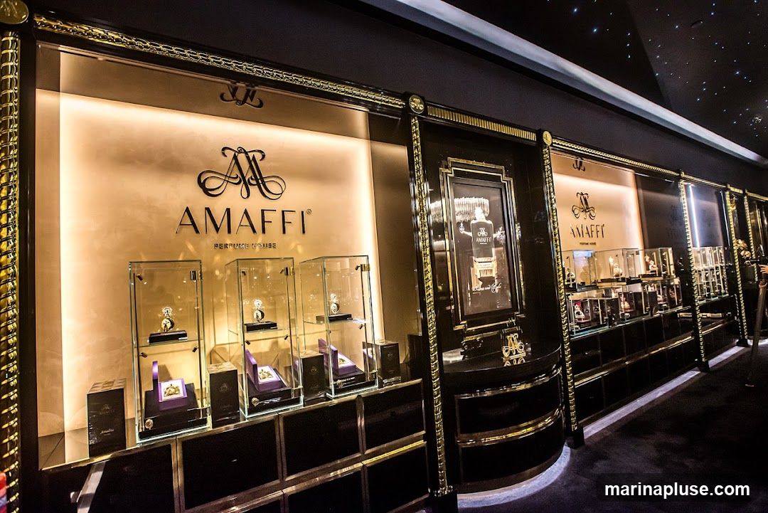 A luxurious interior display for AMaffi Perfume House. The display features a large illuminated sign with the AMaffi logo and name, along with the tagline "PERFUME HOUSE". Several glass display cases showcase perfume bottles and gift boxes. A framed advertisement featuring a perfume bottle is also visible. The overall aesthetic is opulent, with dark, glossy surfaces and gold accents. The ceiling is dark and speckled, resembling a starry night sky.