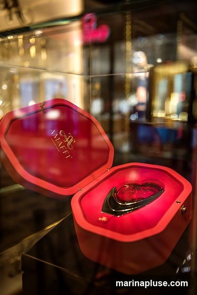 A heart-shaped perfume bottle with a metallic accent is displayed in an open red velvet box. The box is inside a glass display case, with other items visible in the blurred background. The AMAFFI logo is visible on the inside lid of the box.