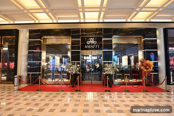 The exterior of a luxury perfume store with a red carpet leading to the entrance. The store features a black marble facade with gold accents and a prominent "AMAFFI" logo above the glass doors. Large floral arrangements flank the entrance, and velvet ropes guide visitors. The interior is visible through the glass, showcasing display shelves with perfume bottles.