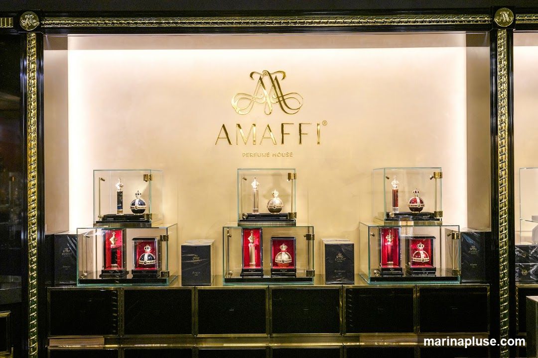 A well-lit display case showcases various perfumes from AMAFFI PERFUME HOUSE. The brand's logo and name are prominently displayed in gold lettering on the wall behind the products. The perfumes are presented in elegant bottles, some resembling crowns, housed in glass display boxes. The overall aesthetic is luxurious and sophisticated, with dark cabinetry and gold accents.