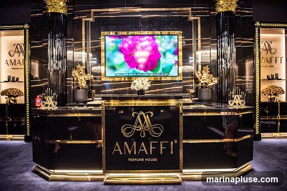 A luxurious display for AMaffi Perfume House features a central counter with the brand logo and name. Behind the counter, a large screen shows a close-up of a pink flower with text overlay. The display is adorned with gold accents, black marble-like surfaces, and decorative gold floral arrangements. To the sides, illuminated display cases showcase perfume bottles.