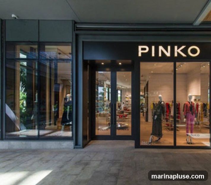 The exterior of a Pinko store with large glass windows and doors. The store name "PINKO" is displayed in large white letters above the entrance. Mannequins dressed in fashionable clothing are visible inside the store.