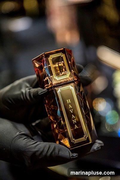 A hand wearing a black glove holds a faceted amber glass perfume bottle with a gold label that reads "HAVANA" vertically. The background is blurred with bokeh lights.