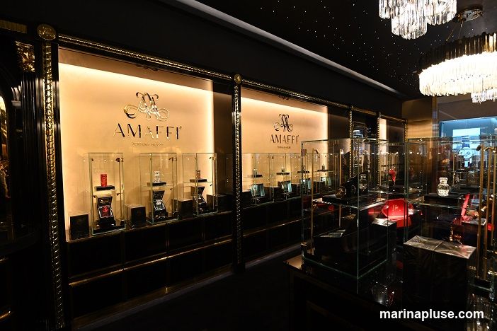 A luxurious interior display of perfumes in glass cases. The brand name 'AMAFFI' is prominently displayed on the wall in gold lettering, along with the tagline 'PERFUME HOUSE'. Various perfume bottles are showcased in illuminated glass cabinets, with some red and black packaging visible. The ceiling features decorative lighting, and the overall ambiance is opulent and sophisticated.