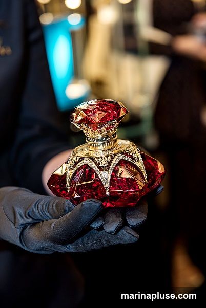 A hand wearing a black glove holds a decorative red perfume bottle with gold accents and a jeweled cap. The background is blurred with warm lighting.