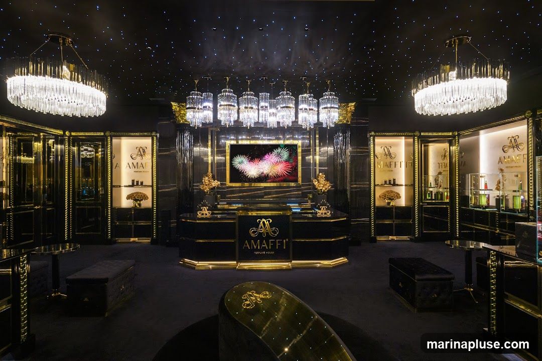 The interior of a high-end perfume store, featuring a black and gold color scheme. The ceiling is adorned with numerous small lights resembling stars, and large, ornate crystal chandeliers hang from above. The store is filled with display cases showcasing various perfume bottles. A central counter with the brand logo "AMAFFI PERFUME HOUSE" is prominently featured, along with a large screen displaying fireworks. Plush seating and decorative elements enhance the opulent atmosphere.