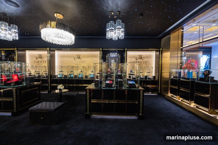 The interior of a high-end jewelry store with a dark, luxurious ambiance. The ceiling is black and adorned with small, star-like lights, complemented by ornate crystal chandeliers. The walls feature illuminated displays showcasing jewelry in glass cases, with the brand name "AMAFFI" prominently displayed. The store has plush dark carpeting and elegant black and gold display cabinets and counters. Seating areas with tufted ottomans are also visible.